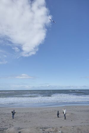 Little boy flying a kite, spending time at the beach with his family, in the background sea with spray and a blue sky with cloudsの写真素材