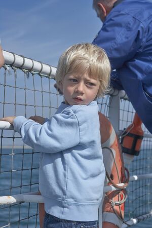Blond Small child clinging to the railing on ferry, looking back for taking a look, in the background a beautiful blue seaの写真素材