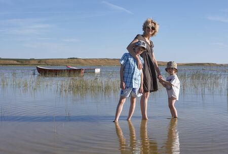 Blond mother with sunglasses, holding the sands of her son while keeping the other one in her arm, walking around in a lake with reed and boats, in Scandinaviaの写真素材