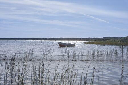 Boat in a blue shimmering sea with aquativ plants and network, in the background a northern danish landscape with houses, and a heaven with a few cloudsの写真素材