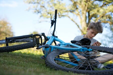 Boy hanging out on a meadow with his smartphone next to his blue bicycle, in the background a treeの写真素材