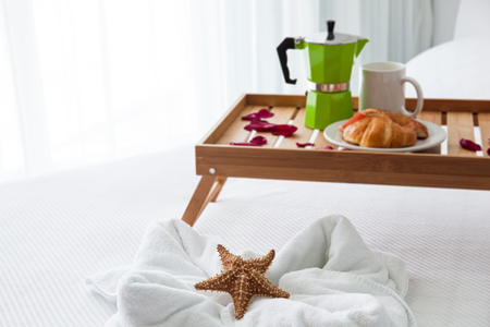 Breakfast wooden tray with coffee percolator and croissant on bed, decorated starfish on white towelの写真素材
