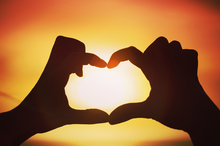 Silhouette of hand in heart shape with sun down in the middleの写真素材