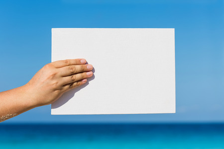 Blank white board in hand against sea view background. Tropical summer vacation conceptの写真素材