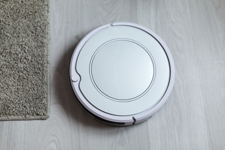 White robotic vacuum cleaner on laminate floor cleaning dust in living room interior. Smart electronic housekeeping technology. Shallow depthの写真素材