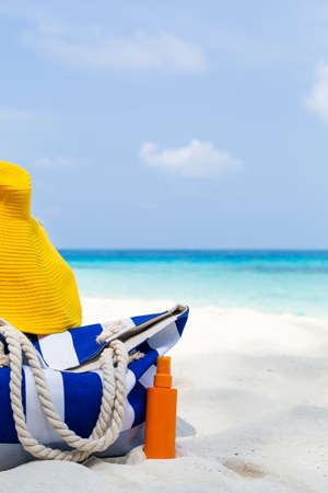Summer hat and blue stripe bag with sunscreen lotion on sandy tropical beach with turquoise sea backgroundの写真素材
