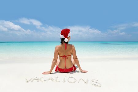 Woman in bikini and Santa hat sitting on paradise beach, enjoying winter holidays and celebrating Christmas at tropical destination. Travel card with perfect getaway for vacationの写真素材