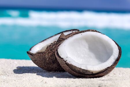 Two halfs of cracked brown coconut on white sandy beach with turquoise sea background, close upの写真素材