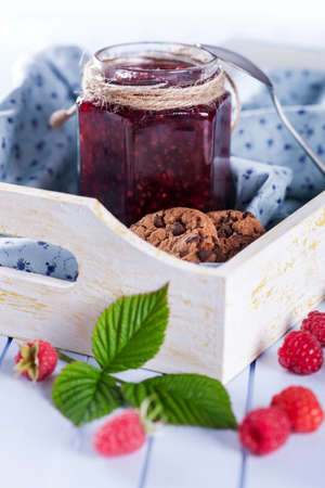 Homemade raspberry jam in jar and tasty cookies in wooden tray decorated with fresh berries and green leaves on table, selective focusの写真素材