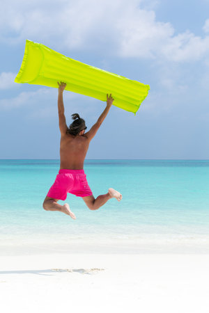 Man with inflatable color green mattress jumping on tropical beach close to sea, enjoying life and...