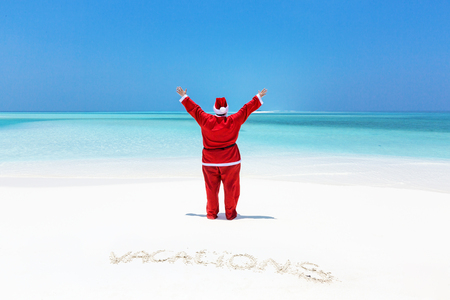 Fat Santa Claus with outstretched arms looking to sea, celebrate Christmas on sandy beach with handwritting word Vacations, enjoy winter holidays at tropical destinationの写真素材