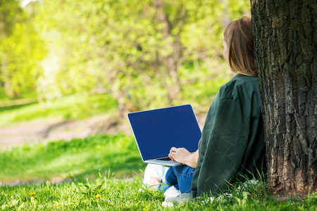 Young blond woman on grass close to tree in summer park using laptop, back view, no faceの写真素材