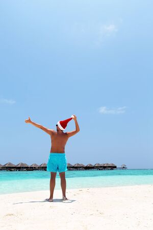 Winter holidays celebration on tropical beach, man on beach in Santa hat with outstreched arms, enjoying Christmas vacation, greeting cardの写真素材