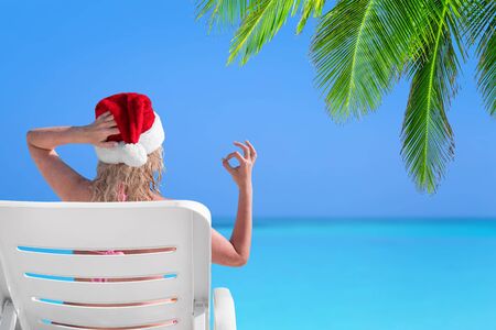 Woman in Santa Claus Hat show gesture Okay on sunbed at beach with palm tree leaf. Christmas vacation on islandsの写真素材