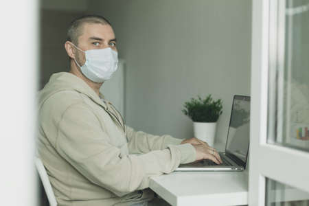 Man in medical mask working at home with laptop, housework quarantine isolation during pandemic coronovirus. Covid-19 conceptの写真素材