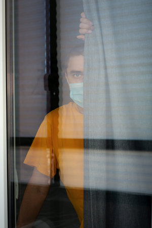 COVID-19 Pandemic Coronavirus. Quarantine man in medical mask on face looking through the window....