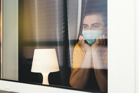 COVID-19 Pandemic Coronavirus. Quarantine man in medical mask on face looking through the window....