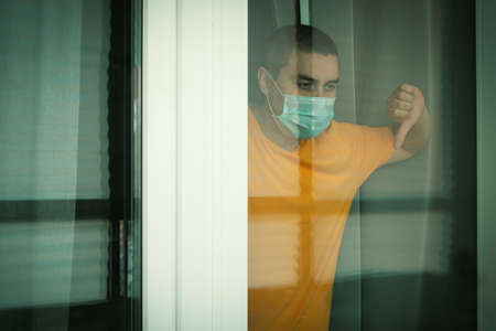 COVID-19 Pandemic Coronavirus. Quarantine man in medical mask on face looking through the window. People on self isolation conceptの写真素材