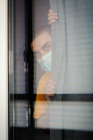 COVID-19 Pandemic Coronavirus. Quarantine man in medical mask on face looking through the window....