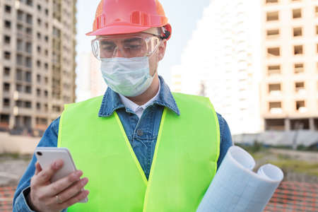 Engineer in red protective helmet and medical mask with blueprint use smartphone at construction site during coronavirus epidemicの写真素材
