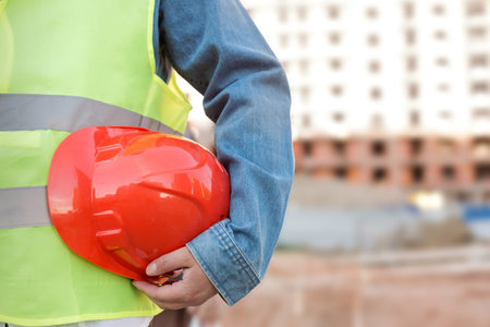 Worker holding red protective helmet at construction site. No face