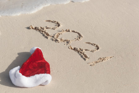 Santa Hat and 2021 number washing away from sand on tropical beach. Christmas celebration