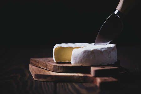 Crop of sliced Camembert with knife on wooden board on dark background, shallow depthの写真素材