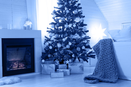 Beautiful decorated glowing fir tree with garland lights near fireplace. Christmas cozy scandic decoration at wooden house. Blue toned imageの写真素材