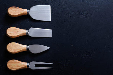 Different types of cheese knives with wooden handles on black background. Top view, menu frame