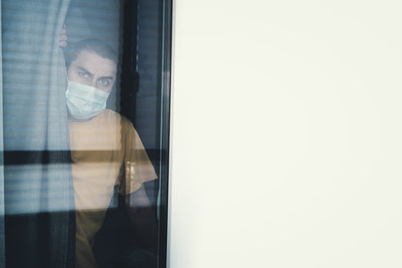 Quarantine man in medical mask on face looking through the window. People on self isolation conceptの写真素材