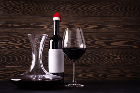 Decanter, glass and bottle wearing Santa Claus hat with red wine on wooden rustic background. Christmas celebration. New year decoration. Closeupの写真素材