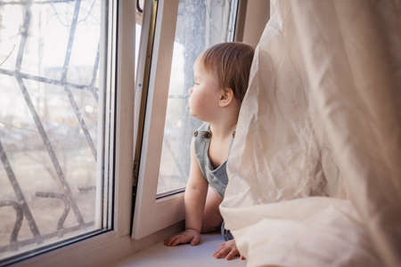 A little child sits in front of the window and looks outside. The windows are closed by the bars. Concept: restriction of movement, isolationのeditorial素材