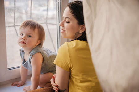 A young woman with a child looking out the window. Window protected by grille, Concept: self-isolation, restriction of movementのeditorial素材