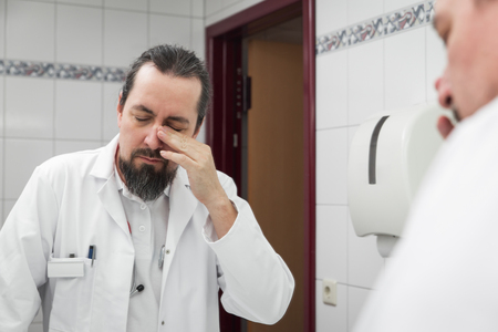 male doctor looks tired into the mirror in a bathroomの写真素材