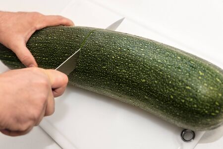 Man is cutting a giant zucchini or courgette on a white board, vegetables in the kitchenの写真素材