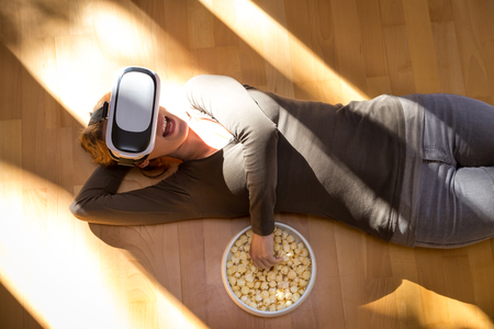 woman is lying with virtual reality glasses on the floor, eating some popcorn, watching a 3d movieの写真素材