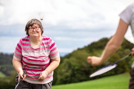 mental disabled woman is playing badminton to train her motor abilities, exercises with a friend or therapist outdoors on a meadow