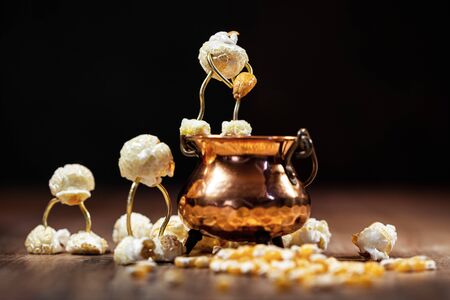 Funny scene with popcorn figures on a wooden table, black background, copper pot with popping maize or corn itの写真素材