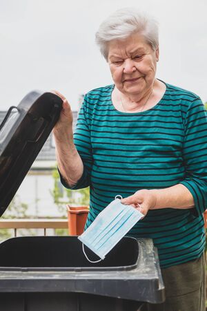 Senior woman holding surgical mask and medical gloves into trash bin, covid-19 and corona pandemic ending and finishingの写真素材
