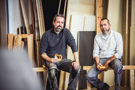 two craftsmen are taking a break in a workshop, two man having a pause while eating a snackの写真素材