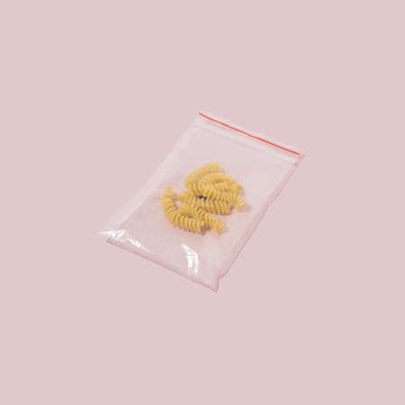 Yellow whole grain spirals in small plastic bag on pastel pink background.  Minimal food concept style.の写真素材