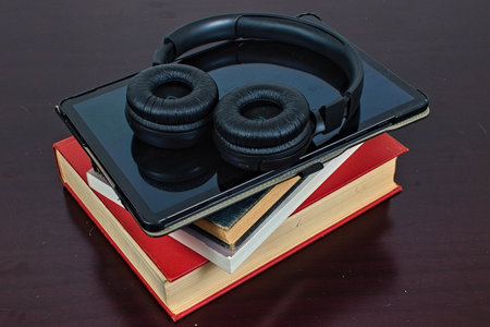 A pair of bluetooth headphones sit on top of a tablet and stack of books. A clash of technology between audiobooks and traditional printed books.の写真素材