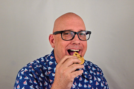 A smiling man enjoys a delicious chocolate chip cookie against a plain backdrop.