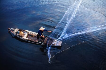 Asian fisherman on wooden boat casting a net for catching freshwater fish in nature river in the early morning before sunriseの写真素材