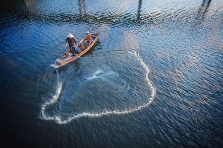 Asian fisherman on wooden boat casting a net for catching freshwater fish in nature river in the...