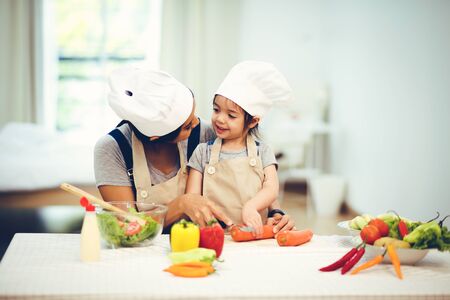 Cute little girl and her mom in chef's hats are cutting vegetables cooking a salad and smilingの写真素材