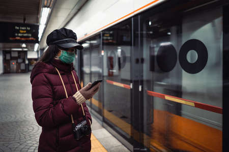 Young Asian woman with protective face mask using smartphone while commuting in the cityの写真素材