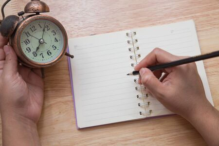 A person are writing messages on blank note book and holding old alarm clock on wooden table vintage toneの写真素材