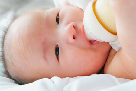 Portrait of a newborn Asian baby on the bed drinks milk from a bottle, Charming black-eyed baby 4 month old lies in bed ,A child resting on a bedの写真素材