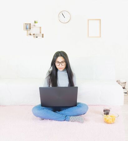 Young beautiful asian woman wore gray t shirt and jean, lying on pink carpet in living room ,work online lifestyle,relax at home with coffee mug,home working laptop,work from homeの写真素材
